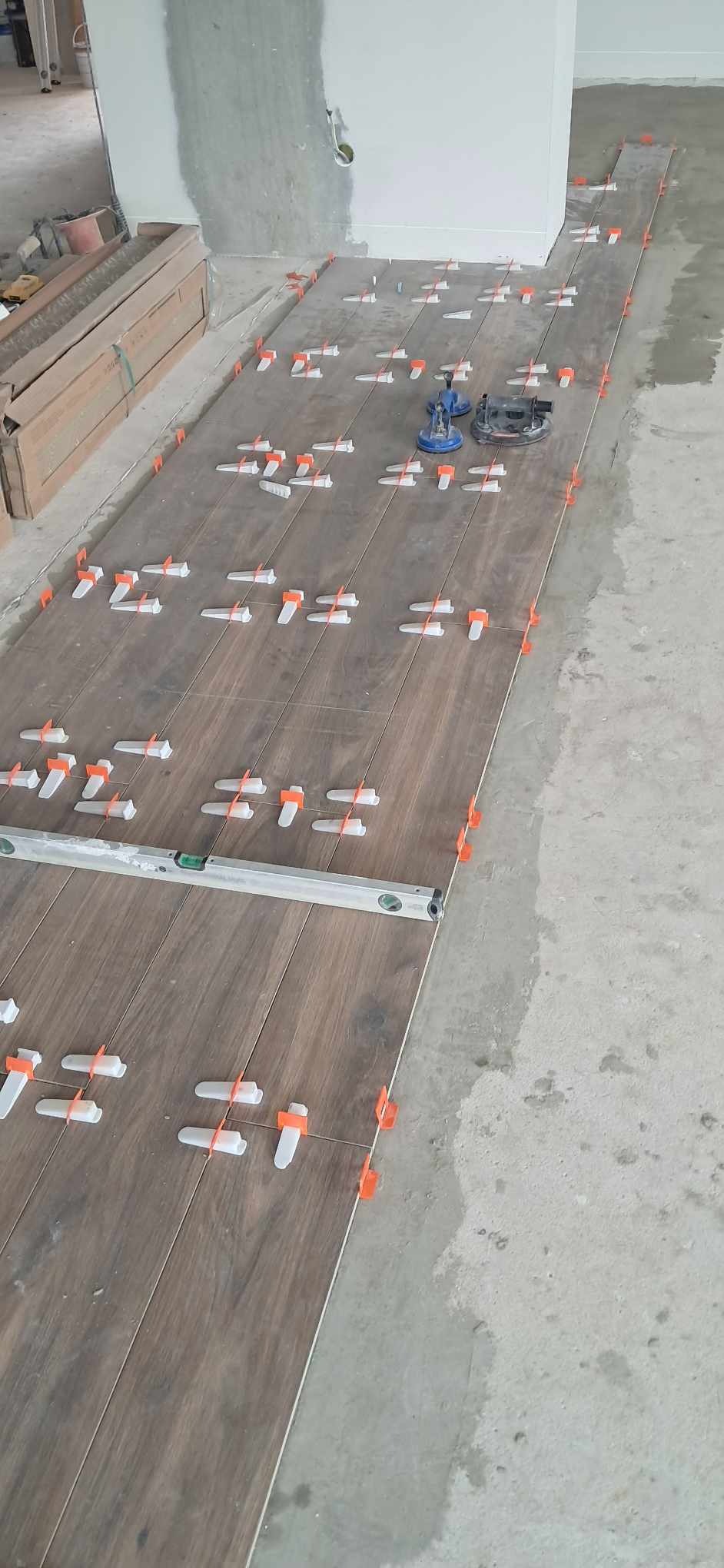Układanie płytek imitujących drewno na podłodze z użyciem poziomicy i systemu poziomowania. Widoczne przyssawki do przenoszenia płytek i krzyżyki dystansowe.