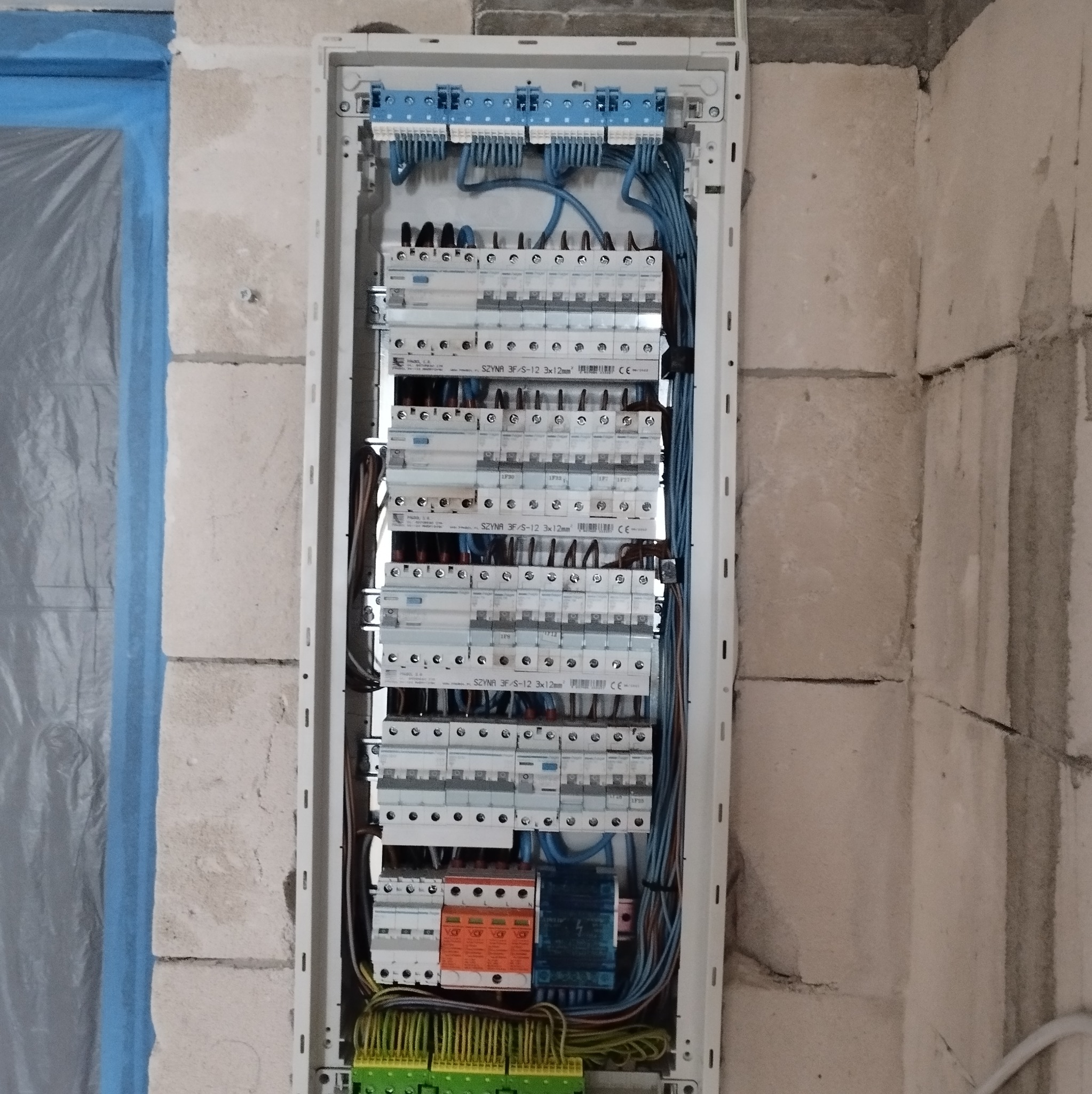 Rozdzielnica elektryczna z zabezpieczeniami, zamontowana na surowej ścianie z betonu komórkowego. Widoczne przewody i listwy zaciskowe. Częściowo zakryte folią malarską.