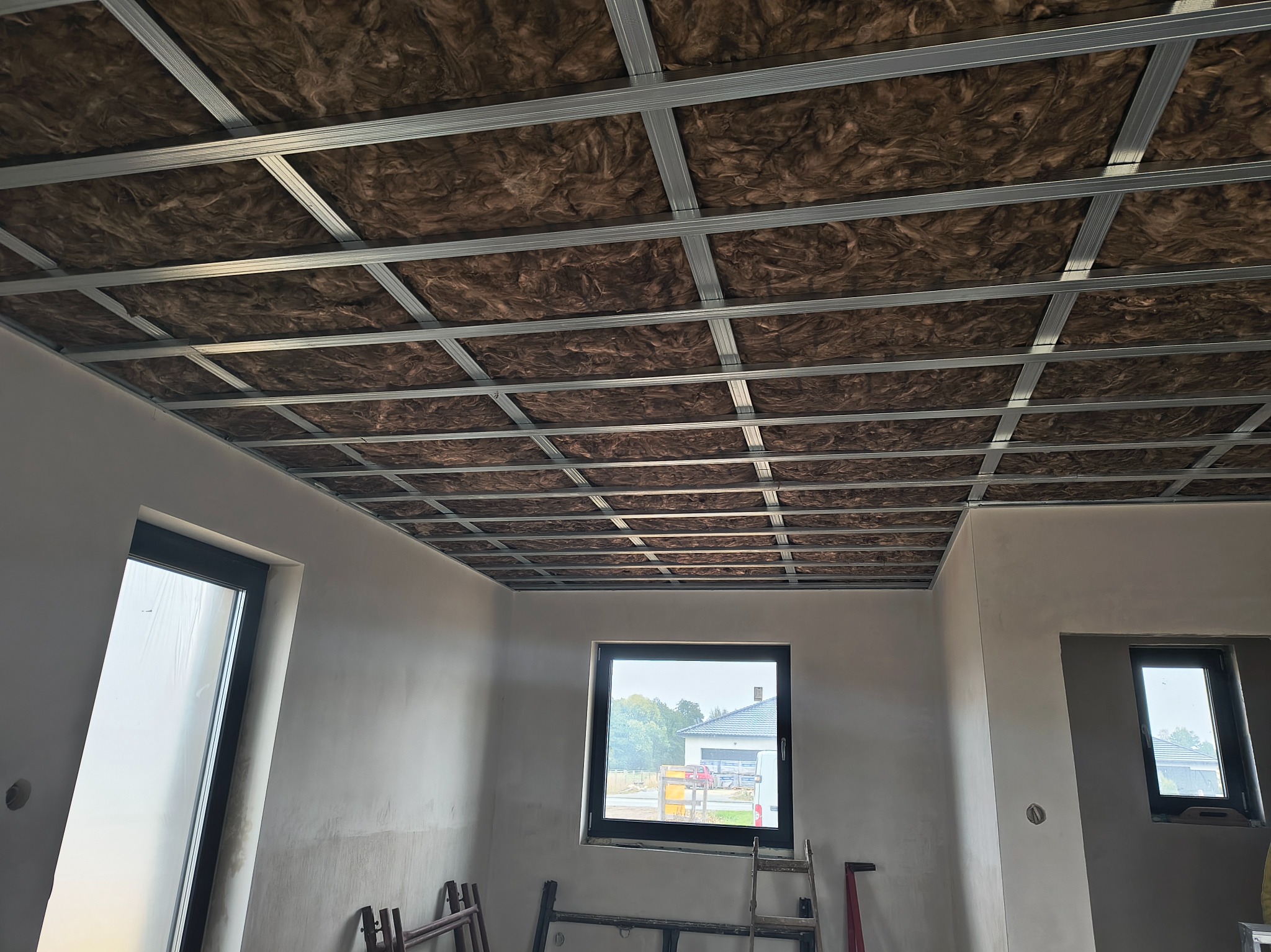 Montaż sufitu podwieszanego z izolacją z wełny mineralnej w jasnym pomieszczeniu z oknami. Widoczna metalowa konstrukcja i drabina. Etap prac wykończeniowych.
