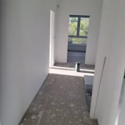 Wnętrze w stanie deweloperskim z świeżo pomalowanymi na biało ścianami, widoczne ślady po pracach na podłodze, oraz narzędzia malarskie w tle.