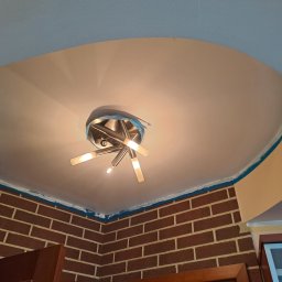 Radosław Bartnowski - Krawędzie sufitu i ściany z cegły zabezpieczone taśmą malarską przed malowaniem, widoczna nowoczesna lampa sufitowa.