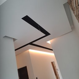 Radosław Bartnowski - Nowoczesny sufit podwieszany z czarnymi elementami dekoracyjnymi i ukrytym oświetleniem LED w jasnym wnętrzu, widok z dołu pod kątem.