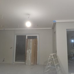 Radosław Bartnowski - Pokój w trakcie remontu: widoczne listwy przypodłogowe z niebieską taśmą malarską, przewody zwisające z sufitu, drabina malarska i zapakowane elementy wykończeniowe oparta o ścianę.