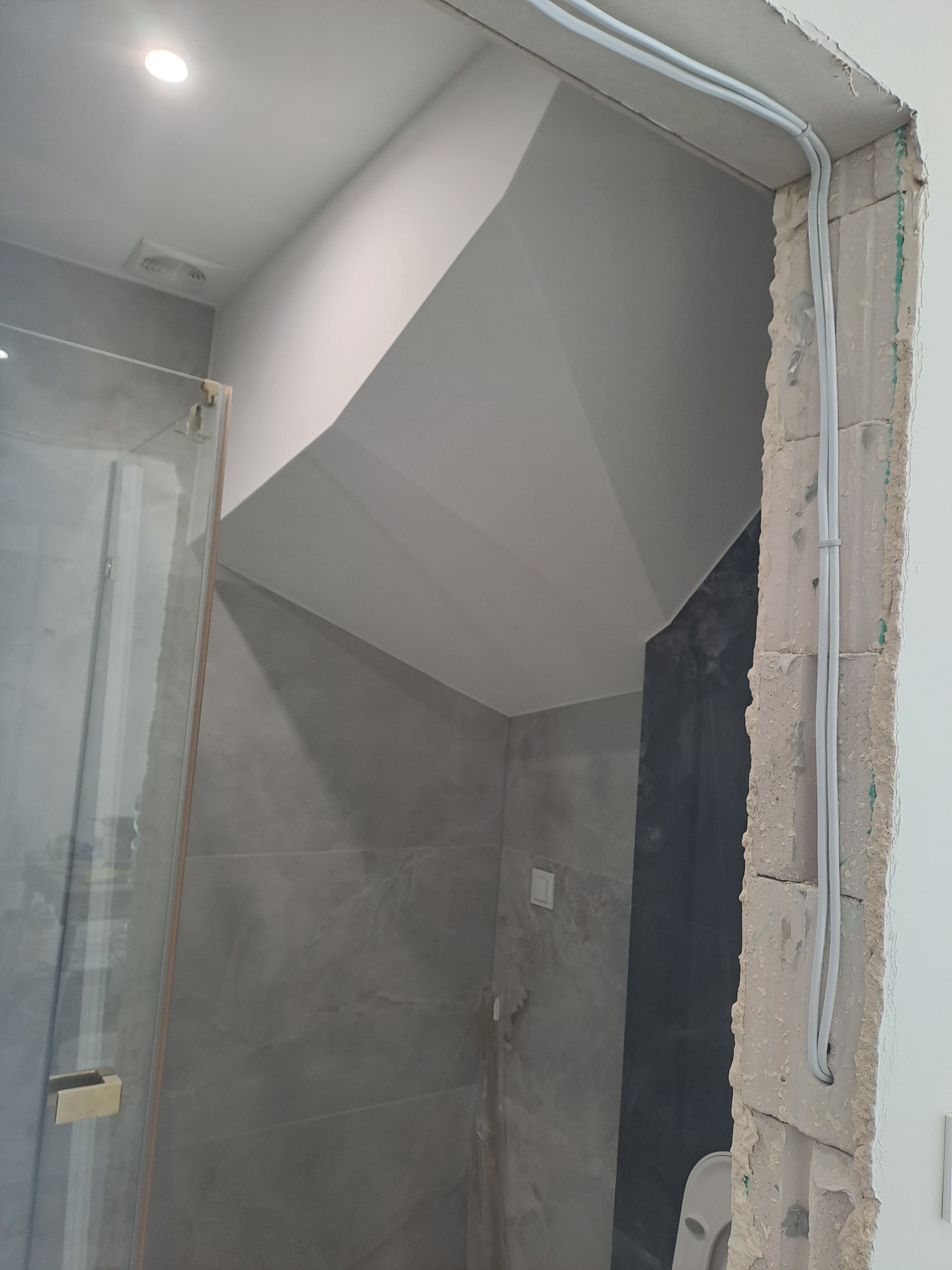 Łazienka w trakcie remontu: widoczne szare płytki, częściowo zamontowana kabina prysznicowa z złotymi okuciami oraz kable elektryczne prowadzone w bruździe w ścianie.