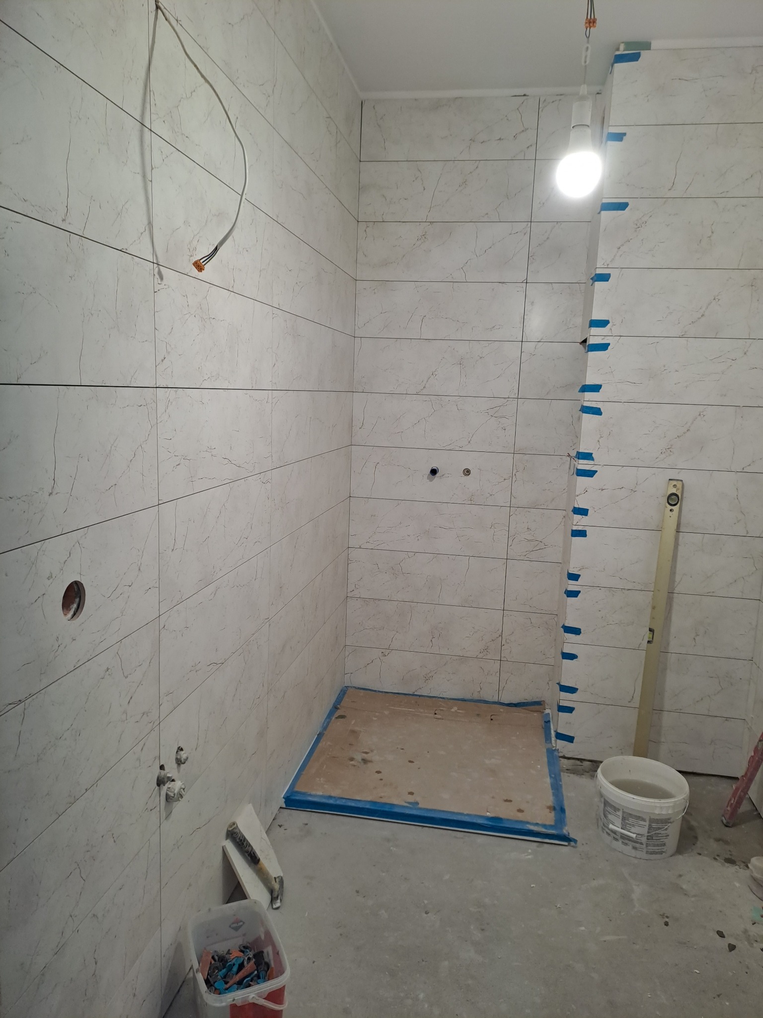 Łazienka w trakcie remontu: ściany wyłożone płytkami imitującymi marmur, widoczne otwory na armaturę, wystające przewody elektryczne, poziomica oparta o ścianę, wiadro z wodą i resztki płytek...