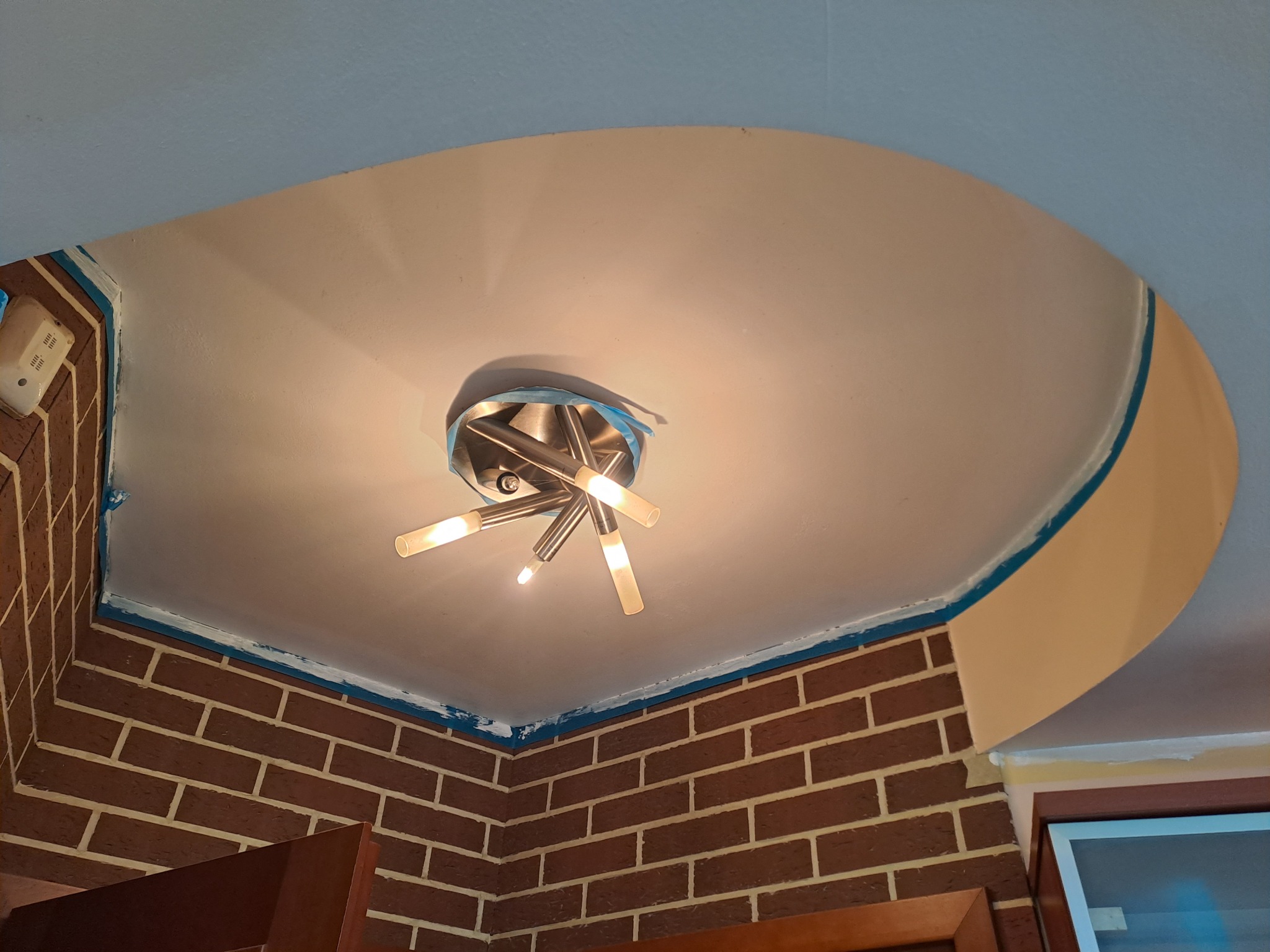 Krawędzie sufitu i ściany z cegły zabezpieczone taśmą malarską przed malowaniem, widoczna nowoczesna lampa sufitowa.