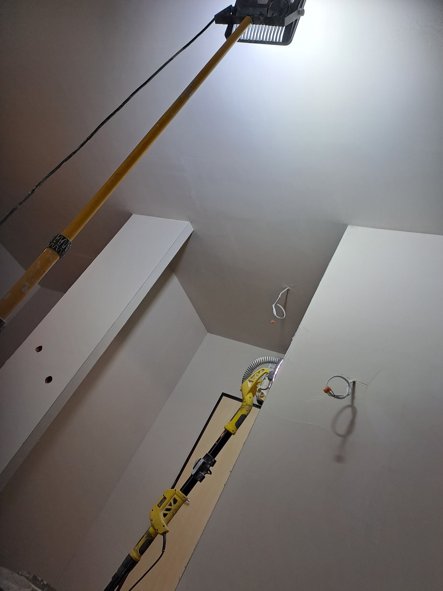 Szlifierka do gipsu na wysięgniku oświetla szlifowany sufit i ścianę z widocznymi puszkami elektrycznymi, w tle fragment ościeżnicy drzwi.