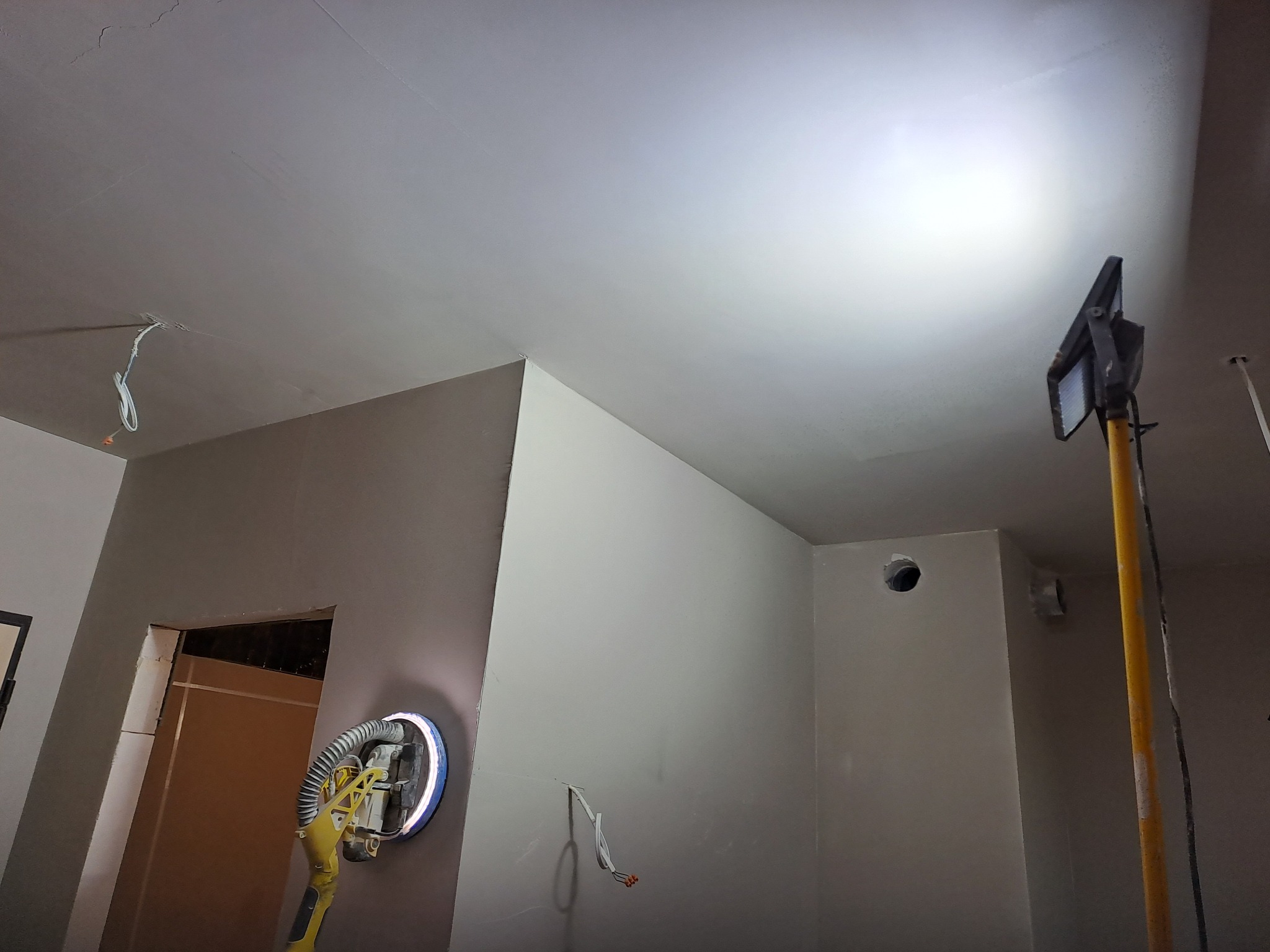 Szlifowanie ściany gipsowej w Pruszczu Gdańskim za pomocą szlifierki z oświetleniem LED, widoczne kable wystające ze ściany i otwór wentylacyjny.