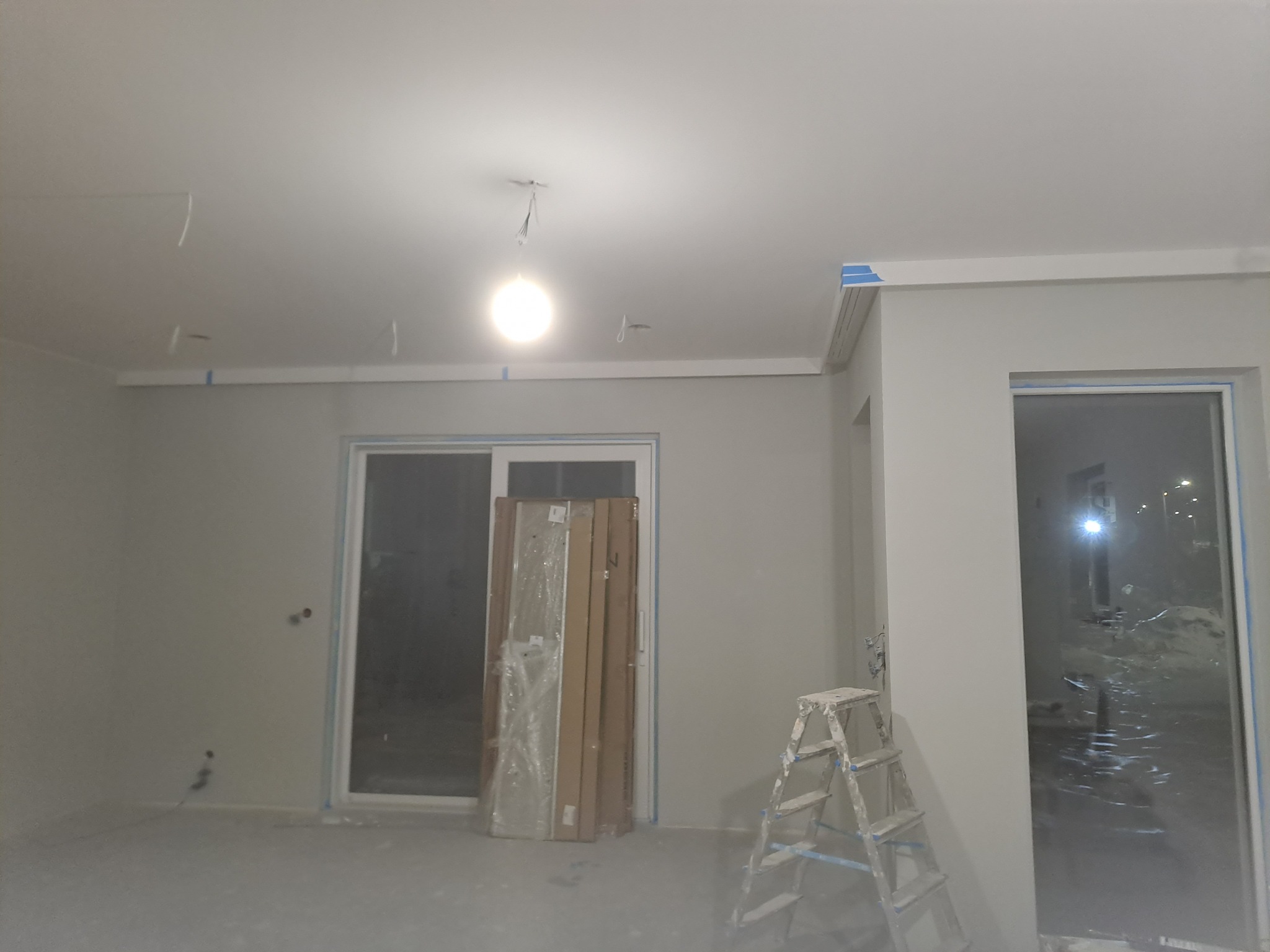 Pokój w trakcie remontu: widoczne listwy przypodłogowe z niebieską taśmą malarską, przewody zwisające z sufitu, drabina malarska i zapakowane elementy wykończeniowe oparta o ścianę.