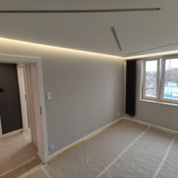 BUDMASTER Ivan Guivan - Pokój po malowaniu ścian na szaro, z podświetlanym sufitem LED, zabezpieczoną podłogą i czarnym grzejnikiem żeberkowym przy oknie.