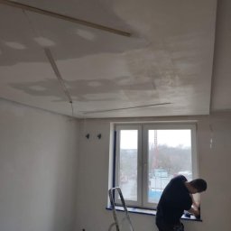 BUDMASTER Ivan Guivan - Mężczyzna na drabinie maluje ramę okienną zabezpieczoną taśmą malarską, widoczny sufit z płyt kartonowo-gipsowych w trakcie szpachlowania, w tle okno z widokiem na miasto.
