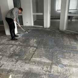 BUDMASTER Ivan Guivan - Mężczyzna maluje betonową podłogę w biurze wałkiem na długim kiju; widoczne ślady wcześniejszych warstw i nierówności podłoża.