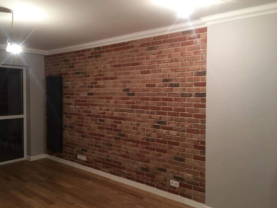 Ściana z cegły dekoracyjnej w pokoju z drewnianą podłogą, grzejnikiem panelowym i białym sufitem z ozdobną listwą.