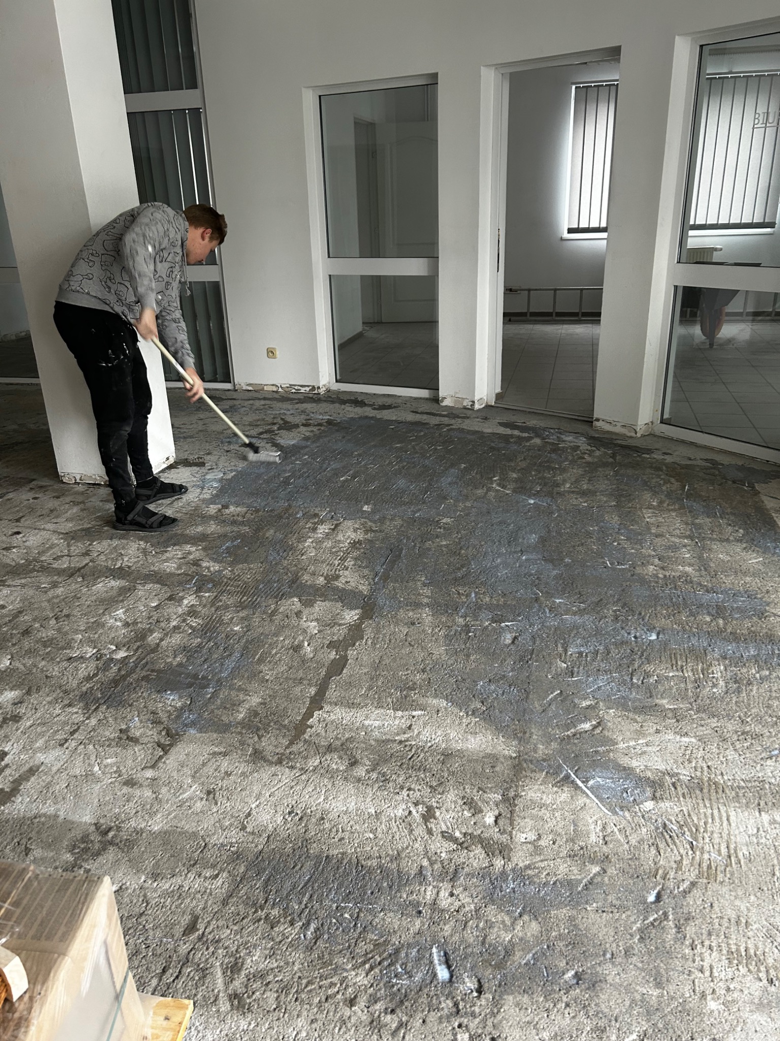 Mężczyzna maluje betonową podłogę w biurze wałkiem na długim kiju; widoczne ślady wcześniejszych warstw i nierówności podłoża.