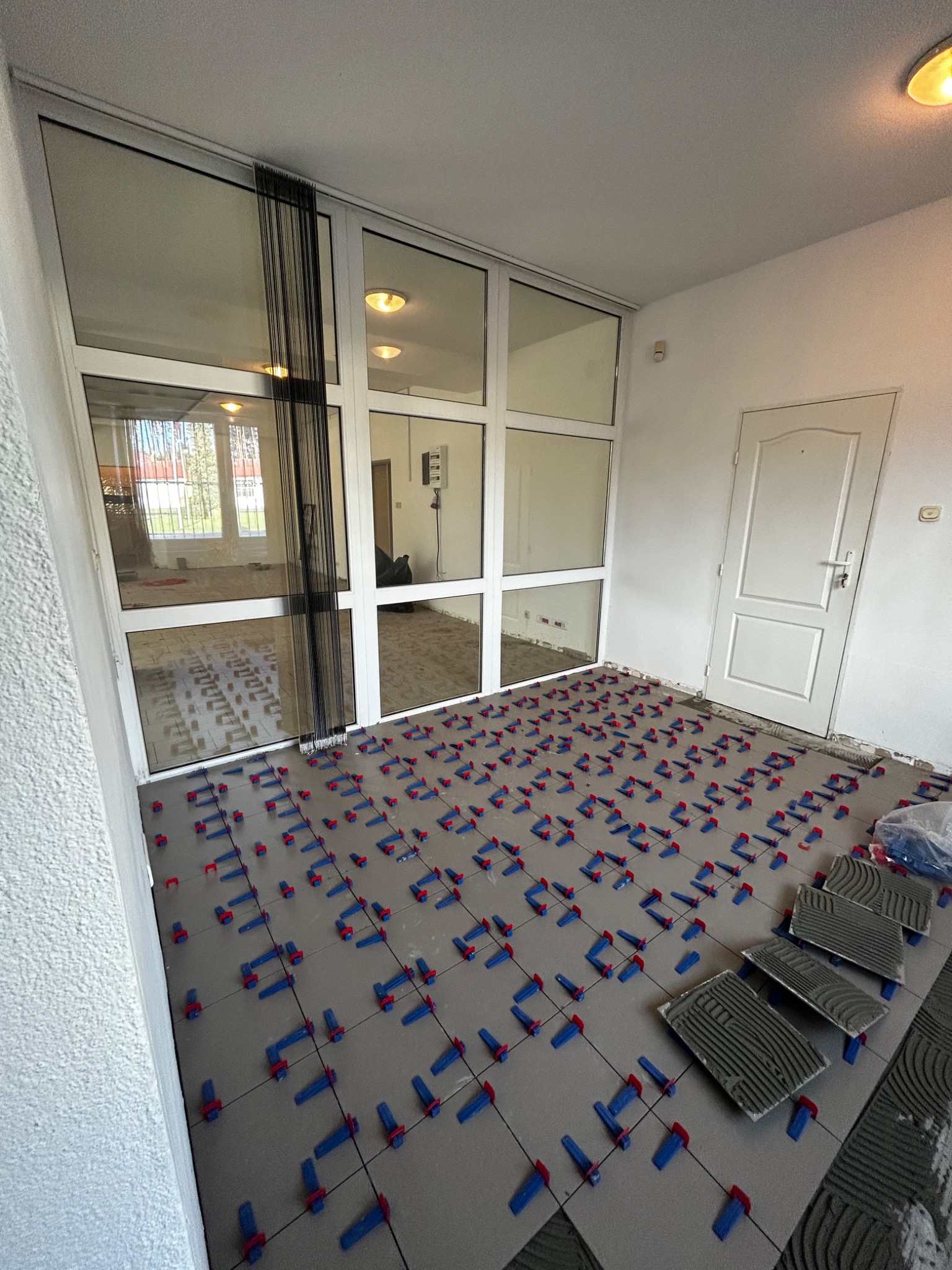 Szare płytki podłogowe w trakcie układania, z zastosowaniem niebieskich i czerwonych klinów poziomujących, widoczne zapasowe płytki z nałożonym klejem oraz duże okno z czarnymi zasłonami w tle.