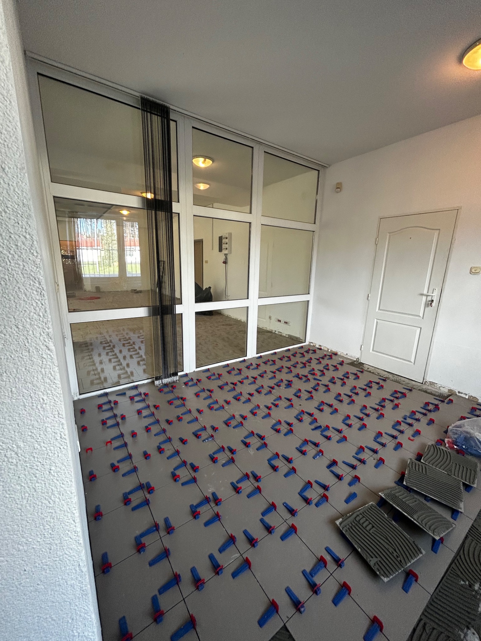 Szare płytki podłogowe ułożone z użyciem systemu poziomowania płytek z niebieskimi klinami i czerwonymi klipsami, widoczne okno balkonowe i drzwi wejściowe.