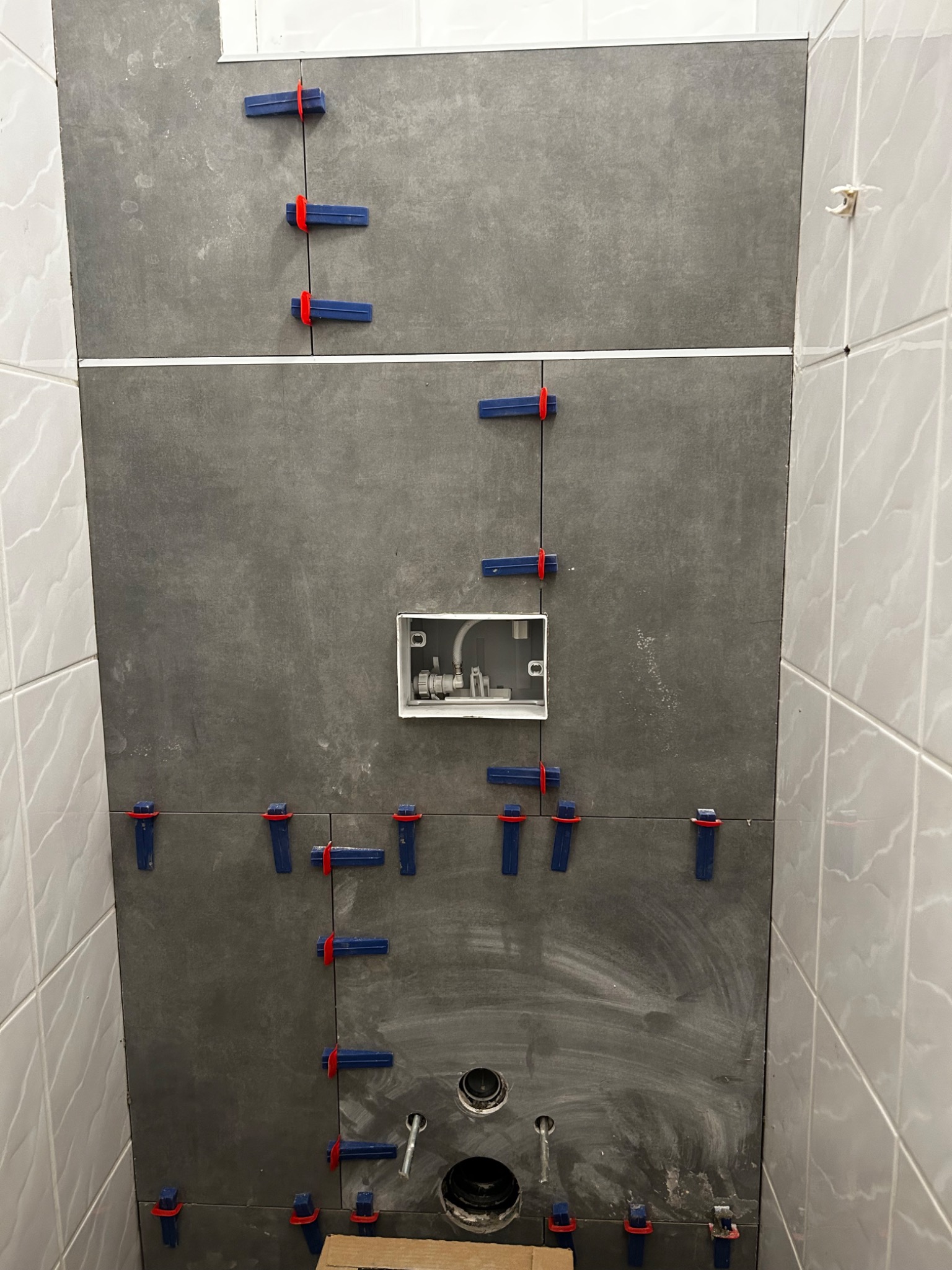 Montaż szarych płytek wielkoformatowych na ścianie w łazience z użyciem klipsów poziomujących, widoczna puszka instalacyjna i otwory na przyłącza kanalizacyjne.