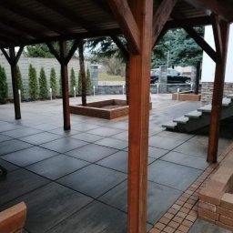 PROJEKT BRUK Paweł Tomczak - Zadaszony taras wyłożony szarą, kwadratową kostką brukową, z drewnianymi słupami podtrzymującymi dach, ceglanymi obrzeżami rabat i schodami prowadzącymi do budynku, w tle rząd tuj i zaparkowany...