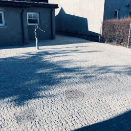 PROJEKT BRUK Paweł Tomczak - Ułożona kostka granitowa w odcieniach szarości, z widocznymi studzienkami kanalizacyjnymi i starą pompą wodną, w tle budynki i fragment ogrodzenia.