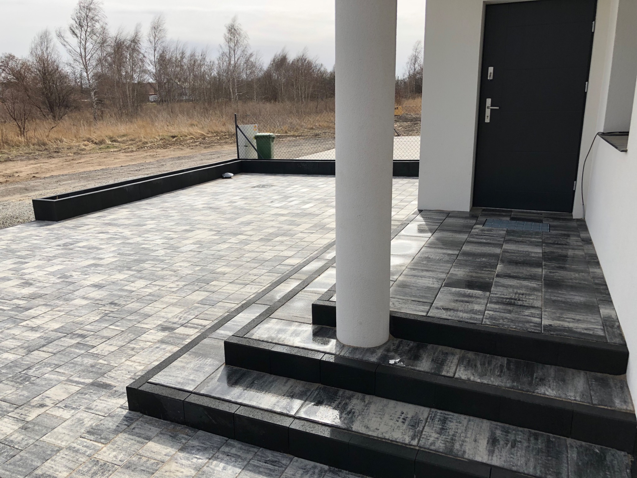 Nowoczesny podjazd i schody wyłożone szarą kostką brukową z czarnymi obrzeżami, prowadzące do wejścia domu z szarą stolarką drzwiową, widok na rabatę i teren za ogrodzeniem.