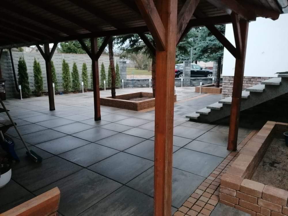 Zadaszony taras wyłożony szarą, kwadratową kostką brukową, z drewnianymi słupami podtrzymującymi dach, ceglanymi obrzeżami rabat i schodami prowadzącymi do budynku, w tle rząd tuj i zaparkowany...