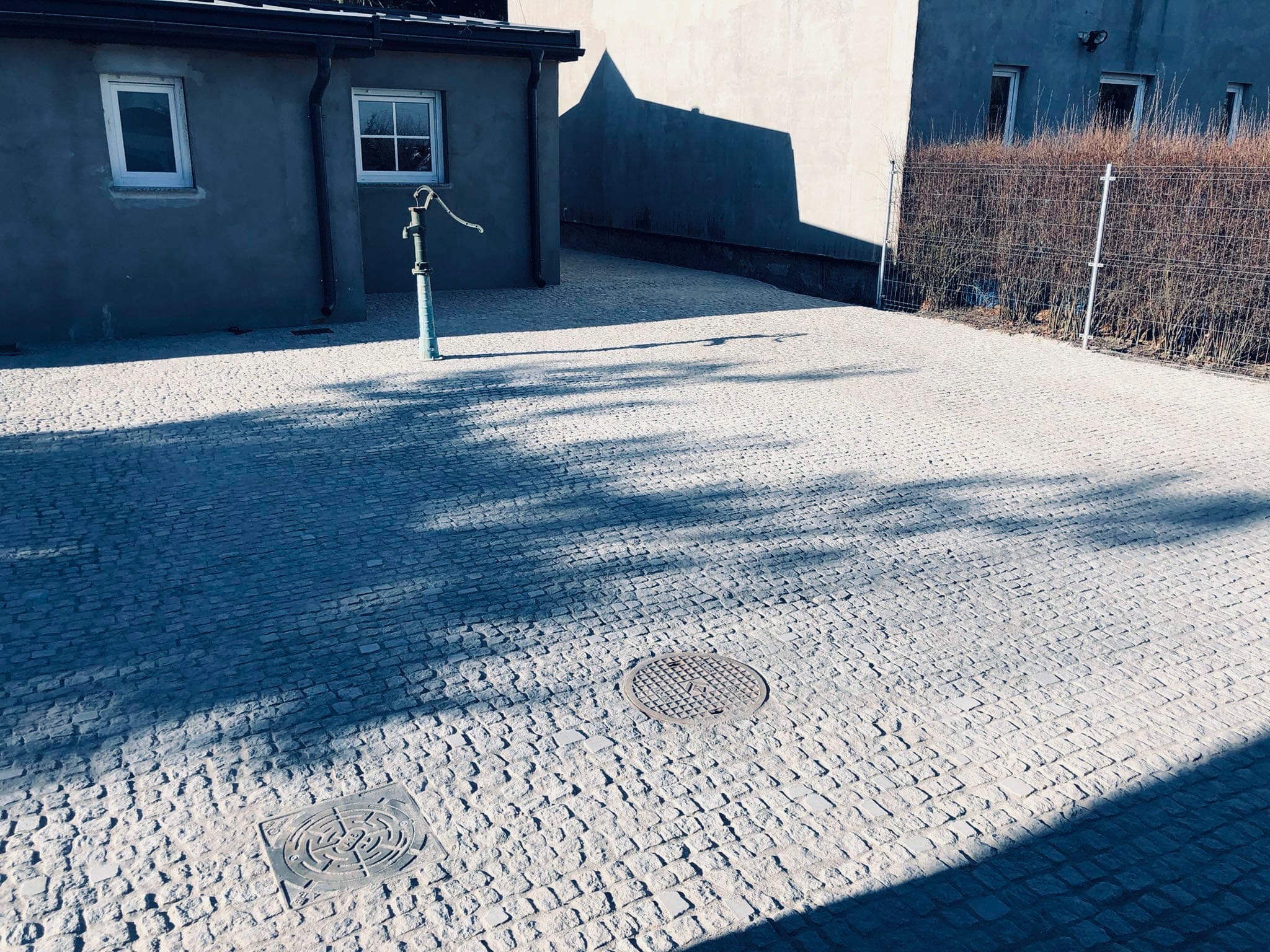 Ułożona kostka granitowa w odcieniach szarości, z widocznymi studzienkami kanalizacyjnymi i starą pompą wodną, w tle budynki i fragment ogrodzenia.