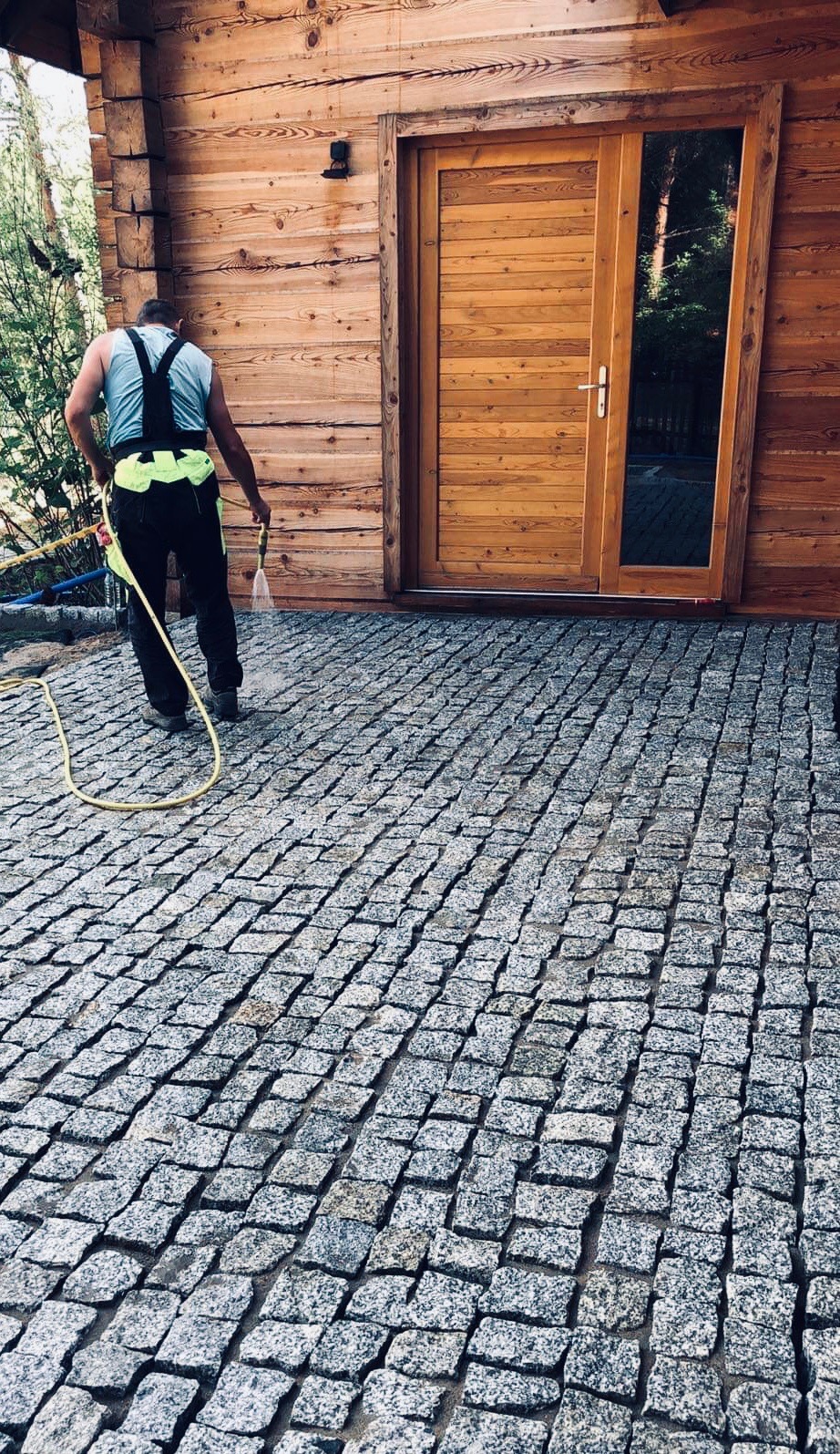 Pracownik w uprzęży myje ciśnieniowo ułożoną kostkę granitową przed drewnianym budynkiem z przeszklonymi drzwiami.