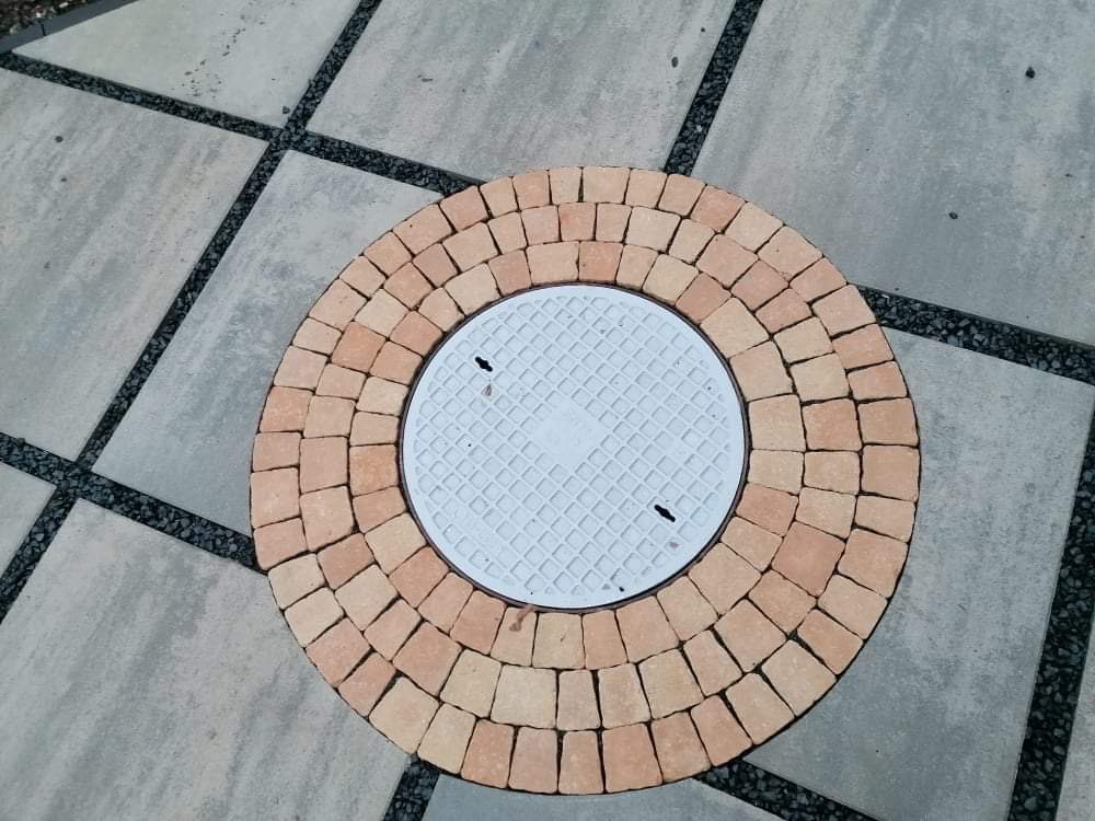 Okrągła studzienka kanalizacyjna z białym, kwadratowym włazem otoczona dekoracyjnym kręgiem z pomarańczowej kostki brukowej, wkomponowana w szarą nawierzchnię z czarnymi fugami.