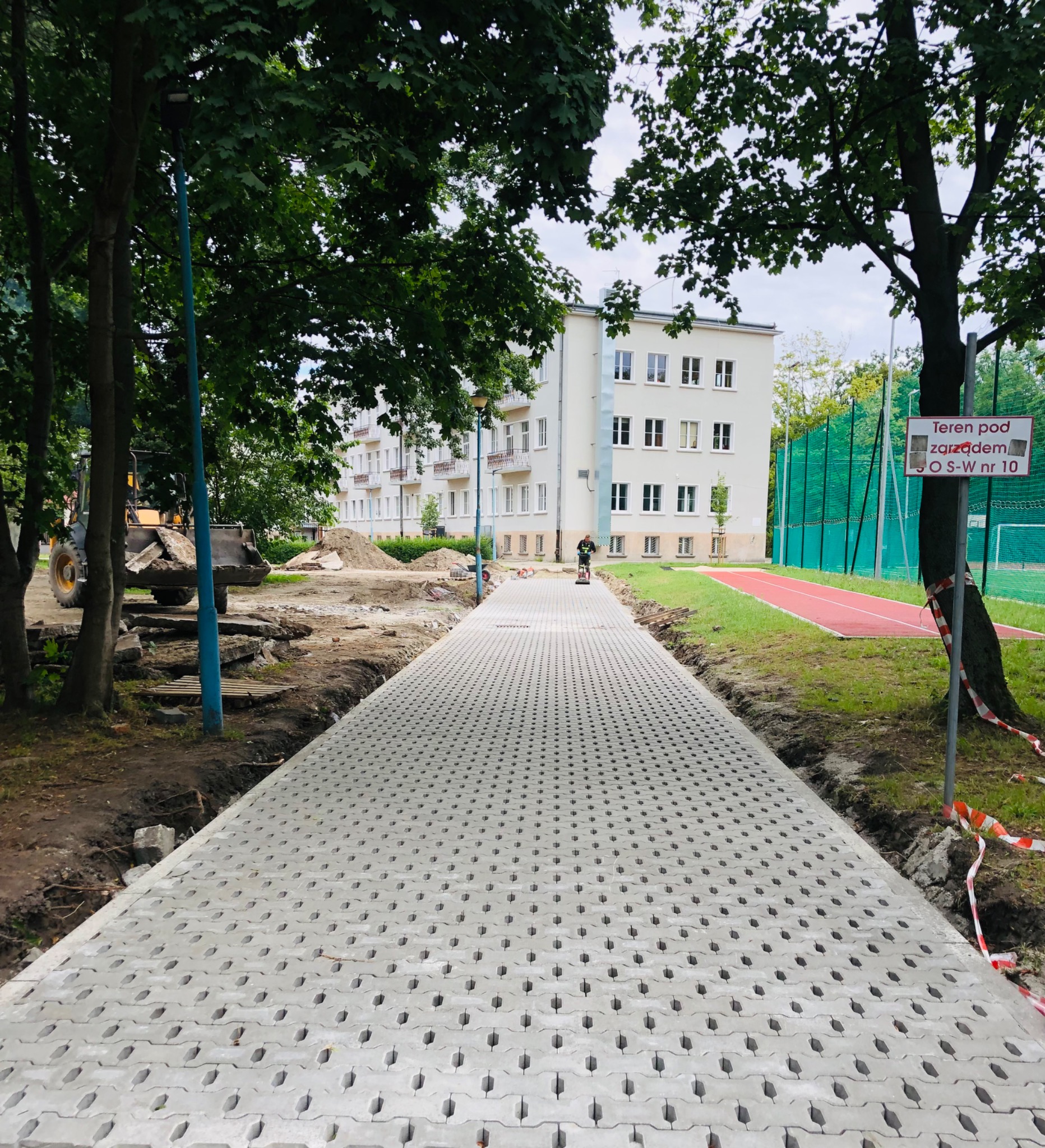 Nowa nawierzchnia z ażurowych płyt betonowych w trakcie układania, widoczny pracownik z zagęszczarką, w tle budynek mieszkalny i teren sportowy.