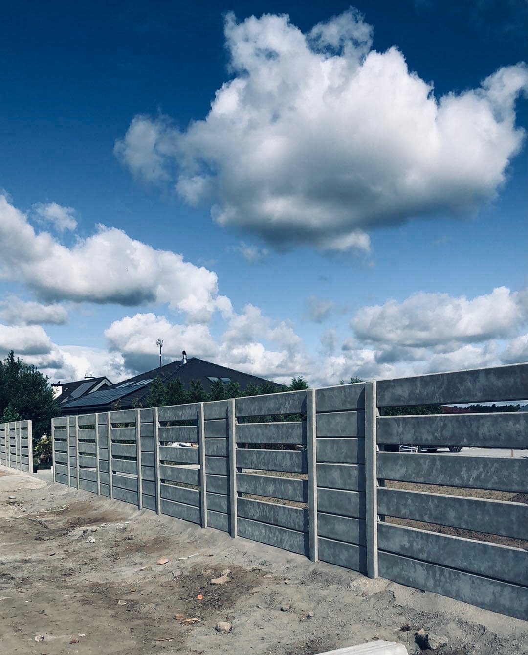 Nowoczesny, betonowy płot panelowy z poziomymi prześwitami, zainstalowany w Tarnowie Podgórnym na nieutwardzonym terenie, z widokiem na dom i błękitne niebo z obłokami.