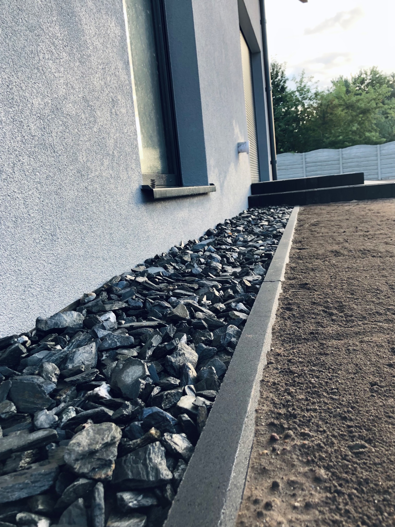 Obsypanie rabaty czarnym kamieniem łupkowym wzdłuż ściany domu z widocznymi schodami i przygotowanym podłożem pod trawnik.