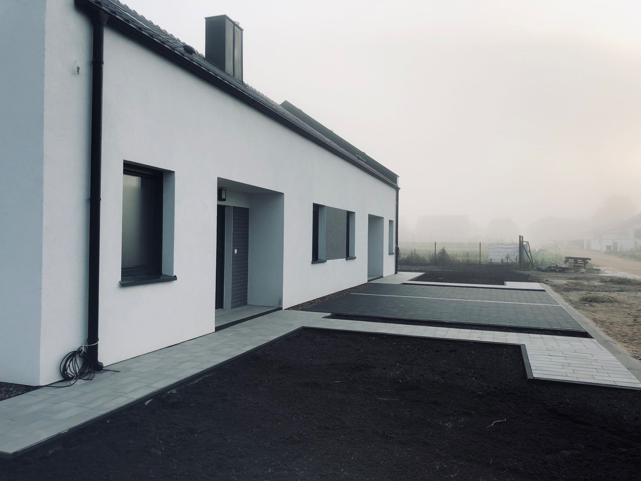 Nowoczesny dom jednorodzinny z białymi ścianami i ciemnymi oknami, z wyłożonym kostką brukową podjazdem i rabatami z czarną ziemią, w tle mgła.