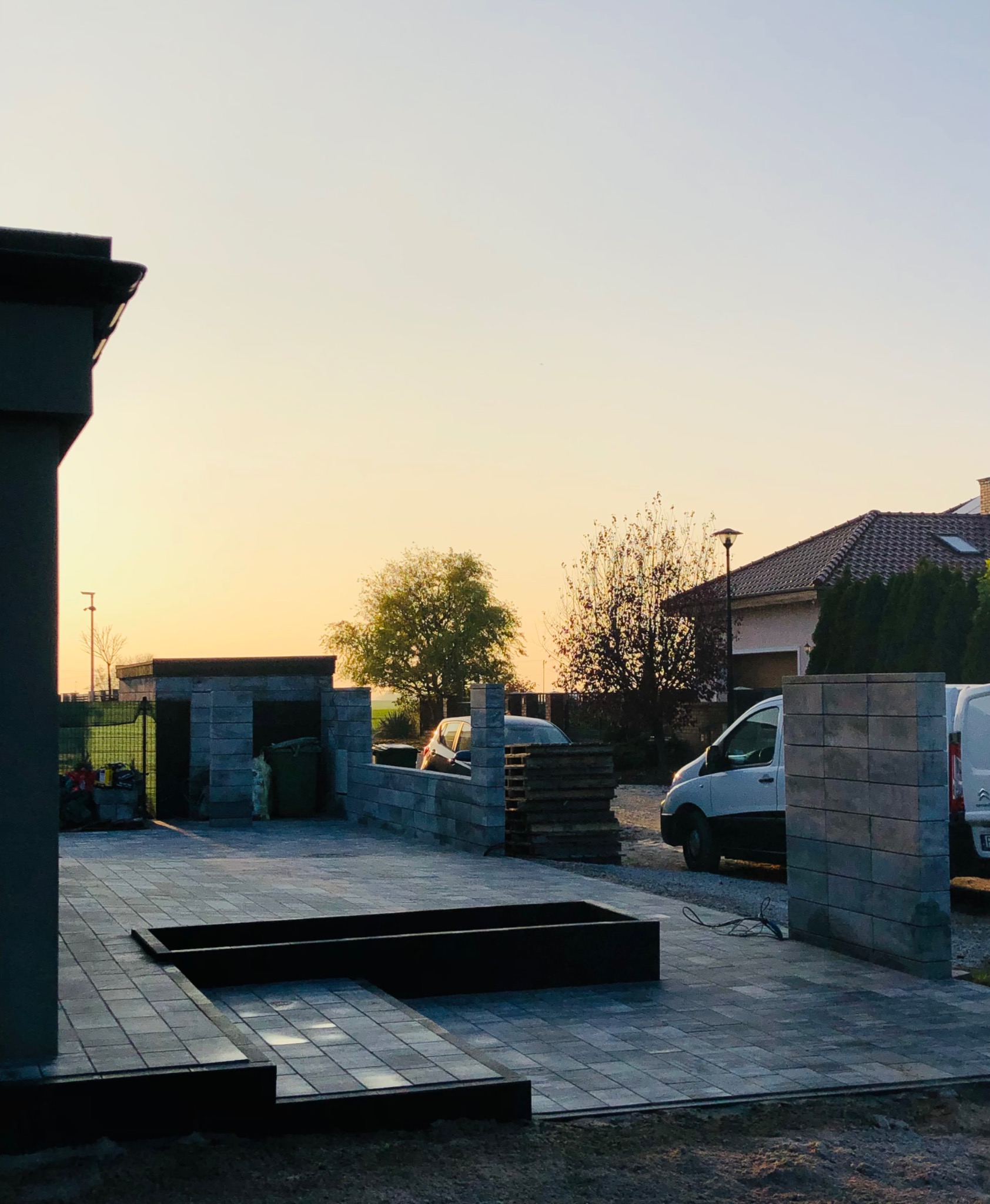 Ułożona kostka brukowa w odcieniach szarości z czarnymi obrzeżami, widoczne elementy małej architektury ogrodowej i fragmenty pojazdów na tle zachodzącego słońca.