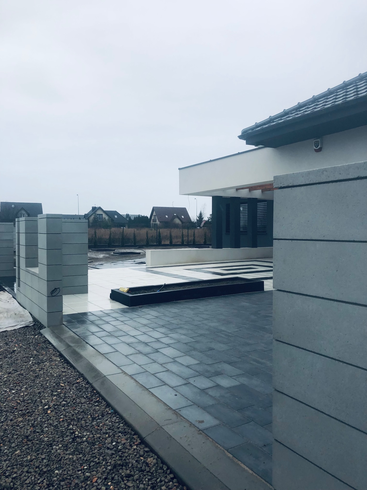 Ułożona, wilgotna kostka brukowa w odcieniach szarości przed nowoczesnym domem z betonowymi elementami ogrodzenia; widoczny fragment posesji z kamienistym podłożem.