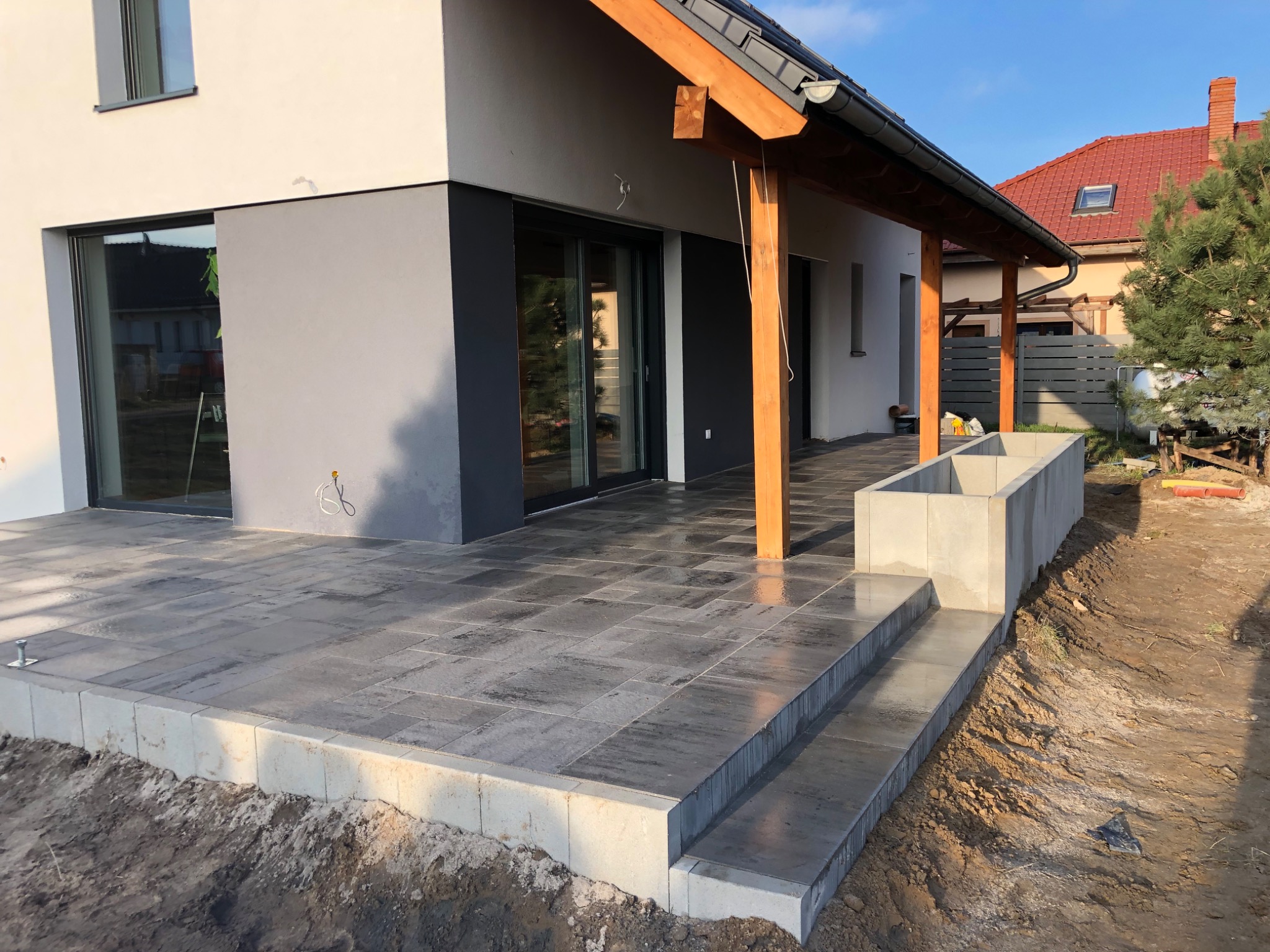 Nowoczesny taras z szarej kostki brukowej ułożony przed domem z drewnianą pergolą i betonową donicą, widoczne schody i świeżo obsypana ziemia.