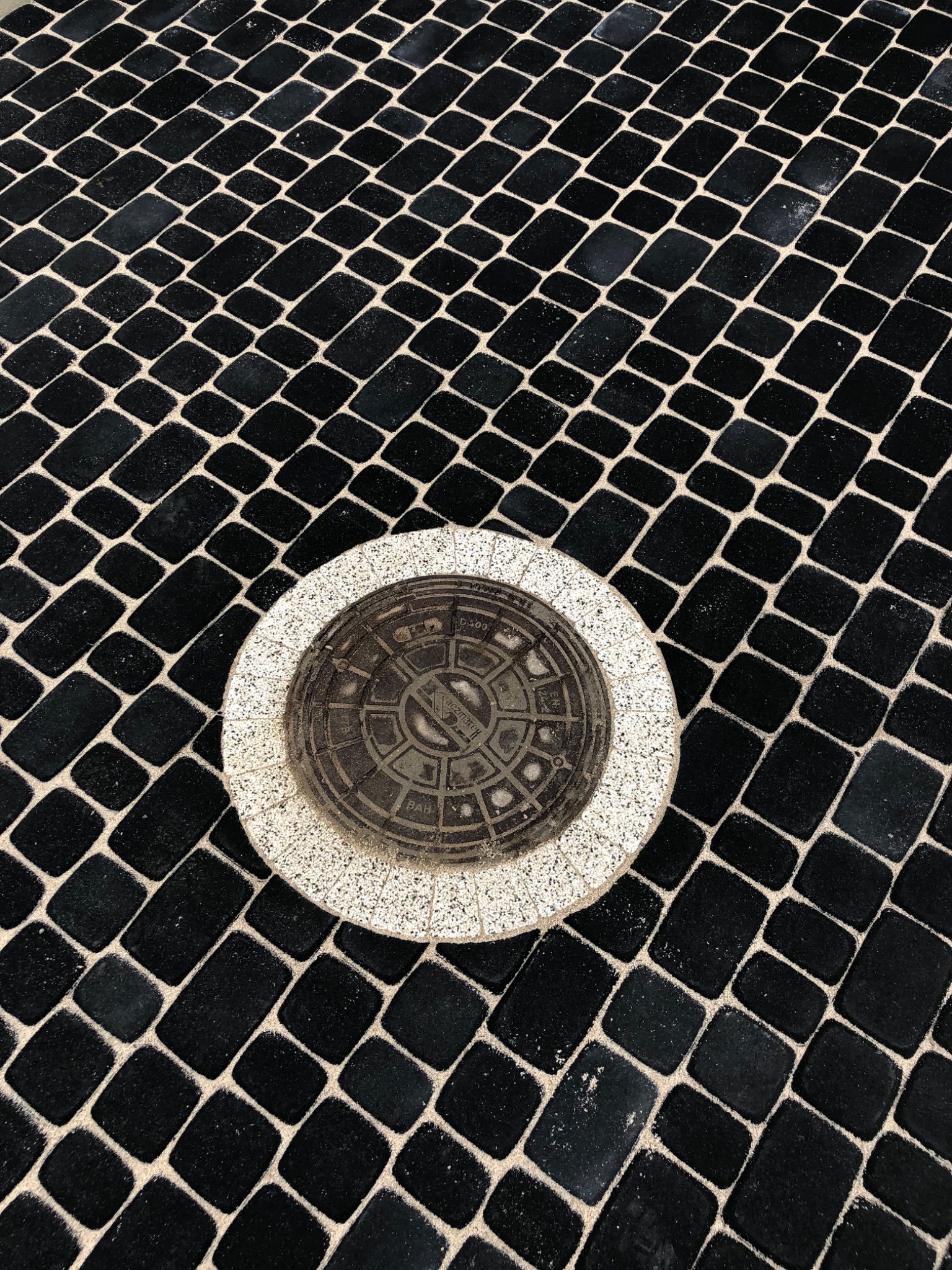 Czarna kostka brukowa ułożona pod kątem z okrągłą studzienką kanalizacyjną otoczoną jasnym brukiem.