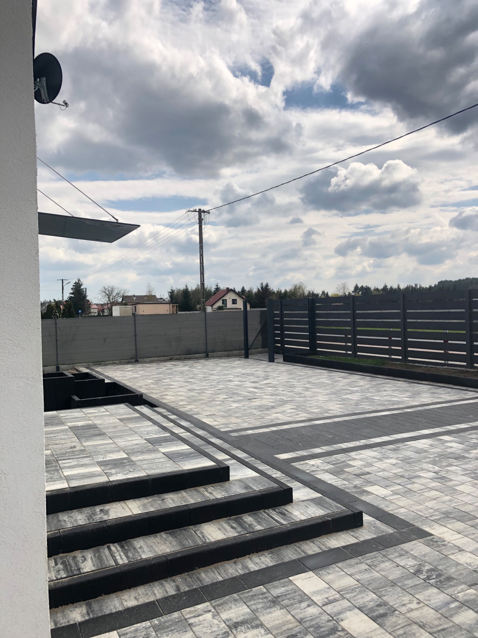 Ułożona kostka brukowa w odcieniach szarości z czarnymi obrzeżami, tworząca wzór na podjeździe i schodach przed domem z szarym ogrodzeniem panelowym w tle.