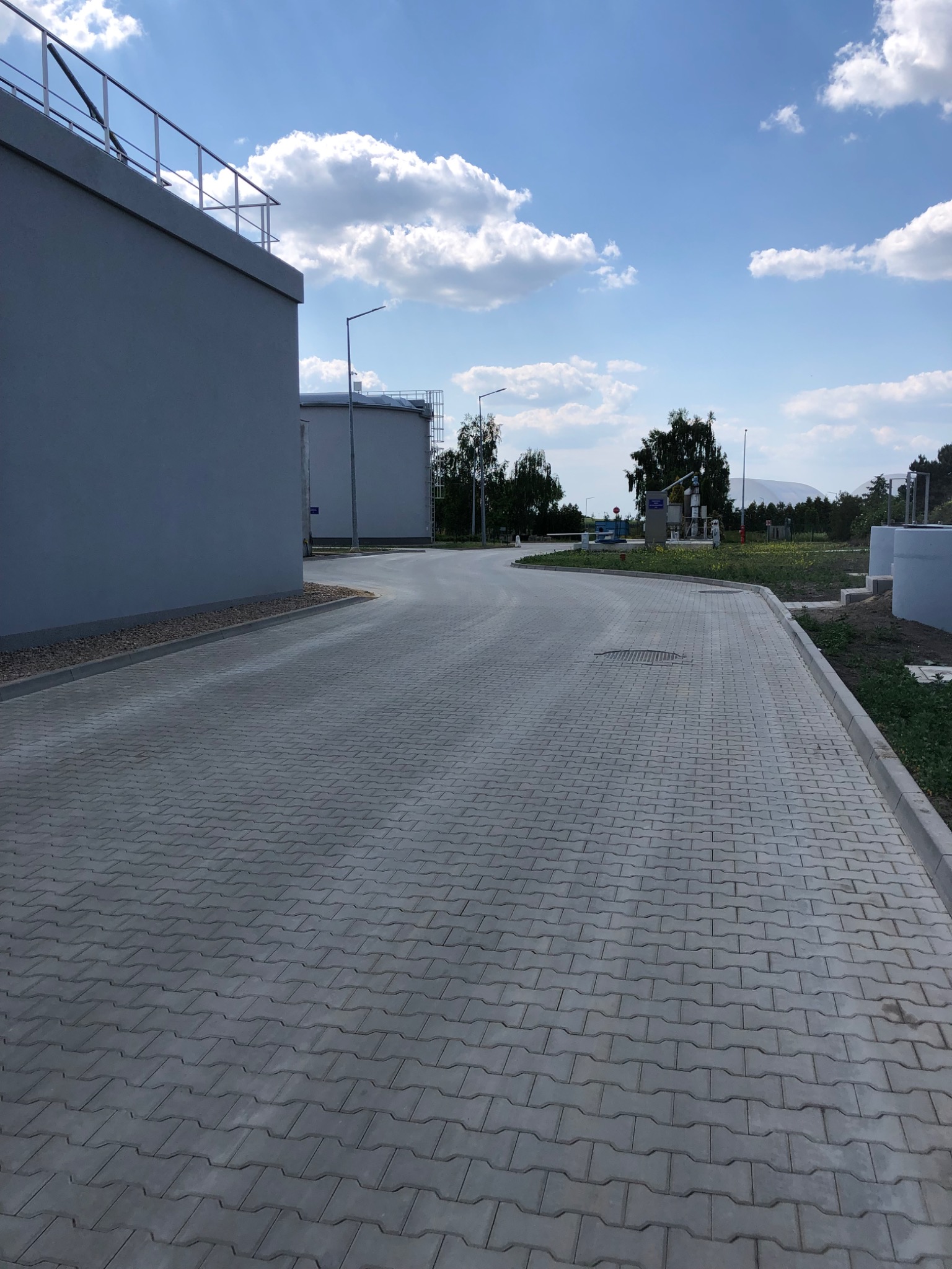 Szeroki, zakręcający chodnik z szarej kostki brukowej prowadzący obok szarych budynków przemysłowych w słoneczny dzień z lekkim zachmurzeniem.