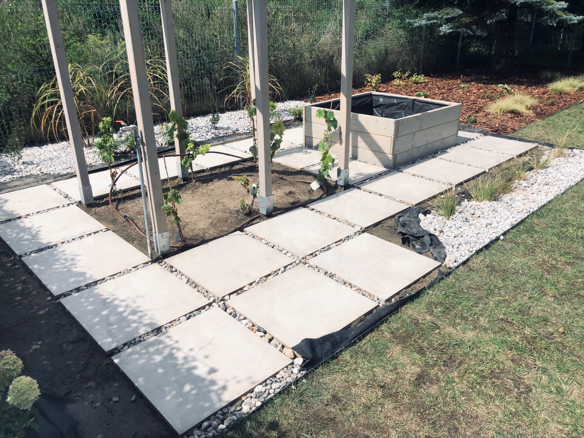 Ścieżka z kwadratowych płyt betonowych oddzielonych białym grysem, prowadząca do drewnianej donicy z czarną wyściółką, widok na ogród z elementami pergoli.