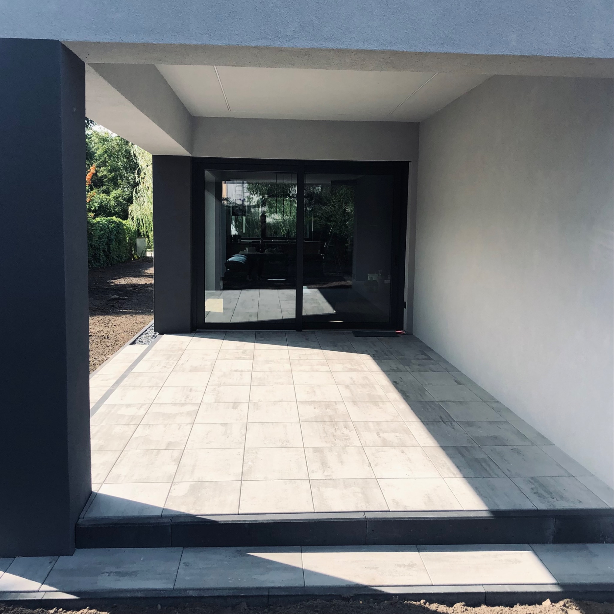 Jasnoszara kostka brukowa ułożona na tarasie przed nowoczesnym domem z przesuwnymi drzwiami tarasowymi, widoczne świeżo obsypane ziemią obrzeża i cień padający na część tarasu.