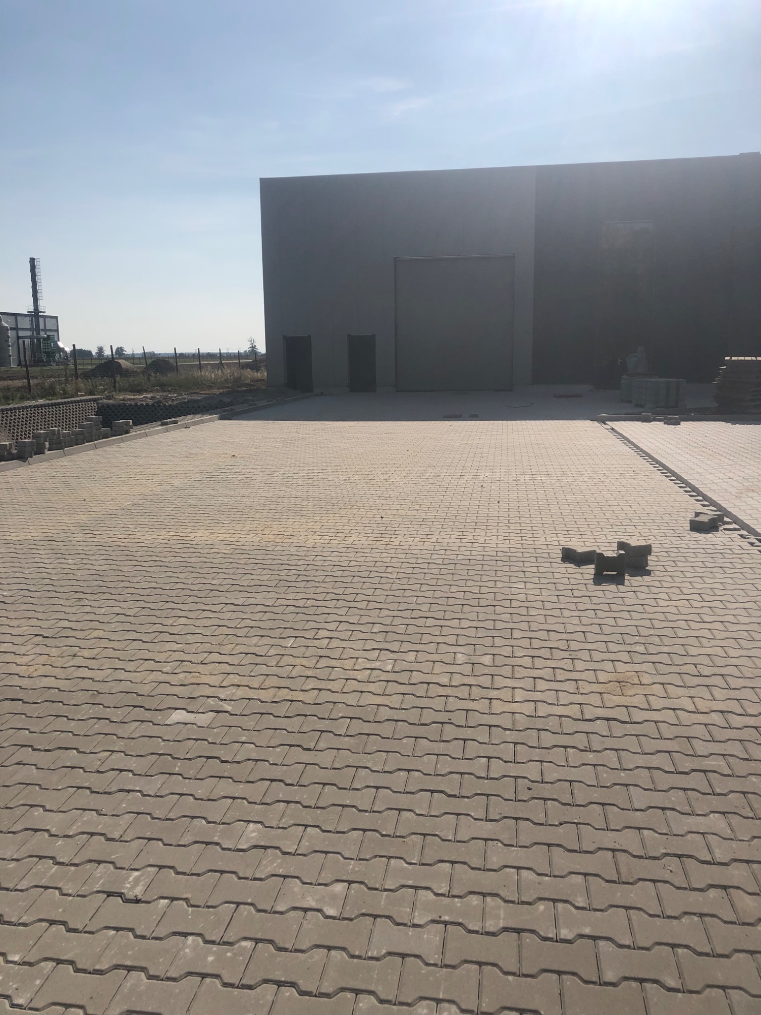 Ułożona szara kostka brukowa przed szarym budynkiem magazynowym, w tle widoczna konstrukcja stalowa i błękitne niebo.