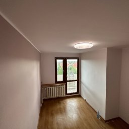 YEVHENII SEMENOV - Pomalowane na jasny róż pokój z drewnianą podłogą i grzejnikiem pod oknem, na zewnątrz widoczna drabina oparta o balkon.