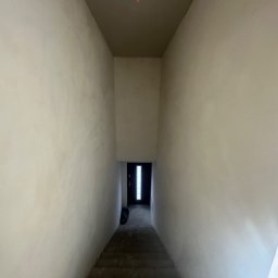 YEVHENII SEMENOV - Korytarz w trakcie wykańczania z widocznymi schodami, surowymi ścianami po szpachlowaniu i przygotowaniu pod malowanie, oraz ciemnymi drzwiami wejściowymi na końcu korytarza.