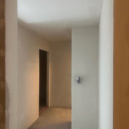 YEVHENII SEMENOV - Korytarz w trakcie prac wykończeniowych, ściany po szpachlowaniu i gruntowaniu, widoczne kable wystające ze ściany zabezpieczone taśmą, surowa podłoga i fragment ościeżnicy przed obróbką.