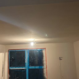 YEVHENII SEMENOV - Przygotowane do malowania pomieszczenie z zabezpieczonymi oknami folią malarską, widoczne przewody elektryczne i gładka, jasna powierzchnia sufitu z delikatnym cieniem.