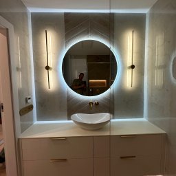 YEVHENII SEMENOV - Nowoczesna łazienka z okrągłym lustrem podświetlanym LED, umywalką nablatową, szafką z szufladami i złotymi akcentami, widoczny fragment podłogi z parkietem ułożonym w jodełkę.