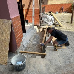 YEVHENII SEMENOV - Pracownik w Szczecinie mierzy odległość przed wylaniem betonu pod schody wejściowe do budynku, widoczne narzędzia i materiały budowlane.