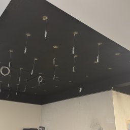 YEVHENII SEMENOV - Czarny sufit podwieszany z punktami świetlnymi w trakcie montażu, widoczne przewody i elementy mocujące. Ściany w surowym stanie, kolumna konstrukcyjna.
