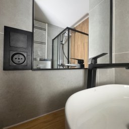 YEVHENII SEMENOV - Nowoczesna łazienka z szarą płytką, czarną armaturą i drewnianym blatem. W lustrze odbija się kabina prysznicowa z drewnianymi panelami. Widoczny kontakt elektryczny.