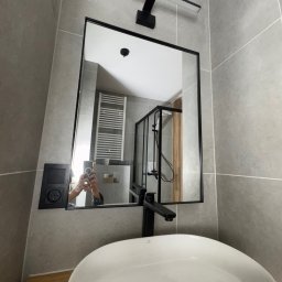 YEVHENII SEMENOV - Nowoczesna łazienka z umywalką nablatową, czarną armaturą i lustrem w czarnej ramie. W tle widoczny grzejnik drabinkowy i kabina prysznicowa.